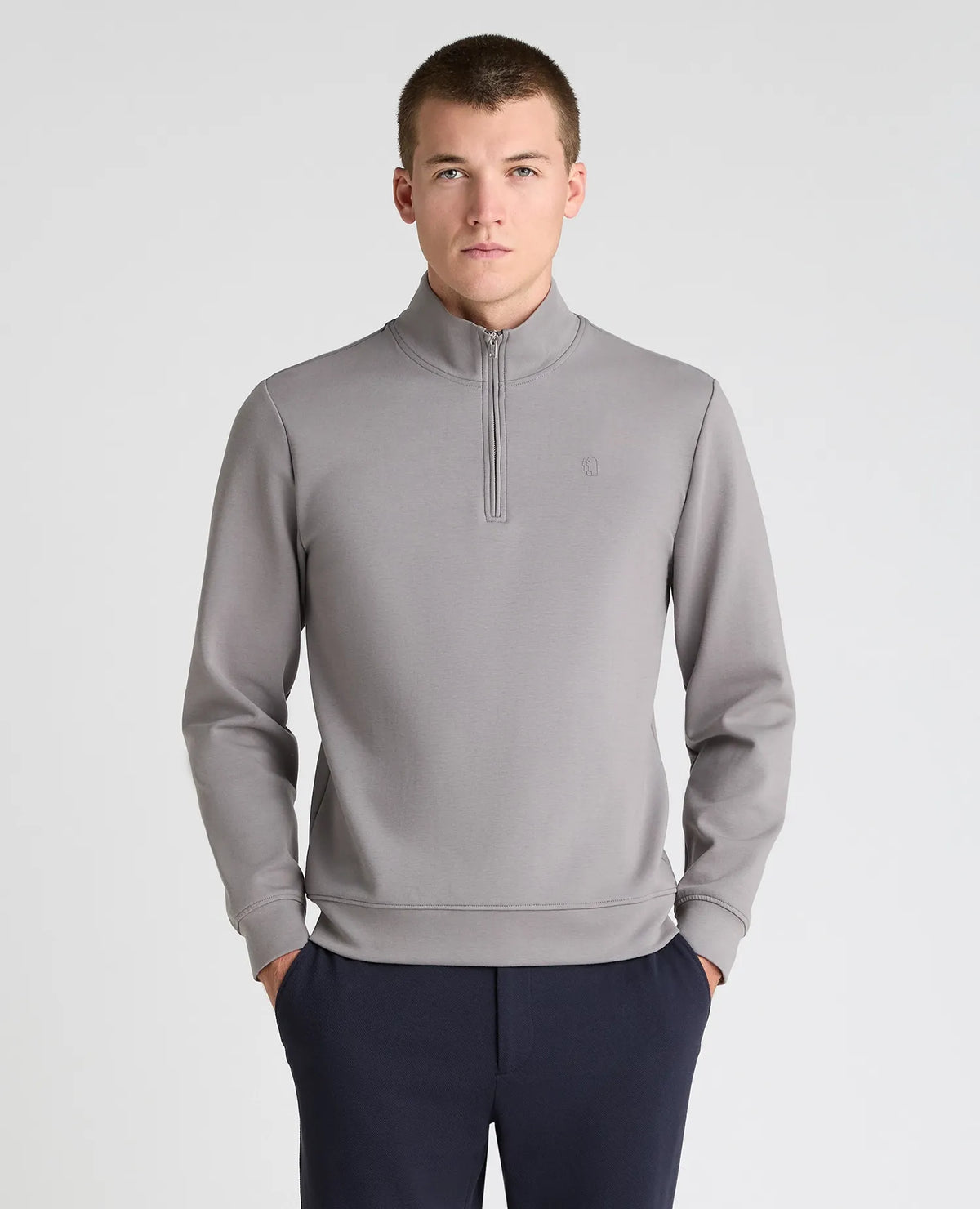 A man wearing a light gray quarter-zip pullover sweatshirt and navy blue trousers, standing against a light gray studio background.
