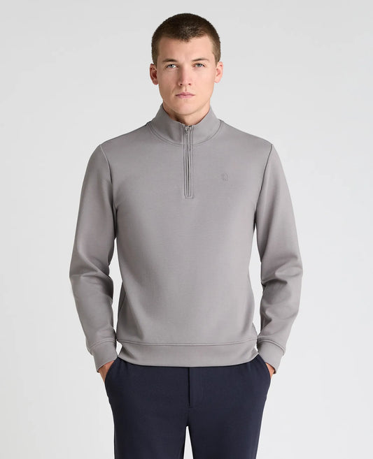 A man wearing a light gray quarter-zip pullover sweatshirt and navy blue trousers, standing against a light gray studio background.