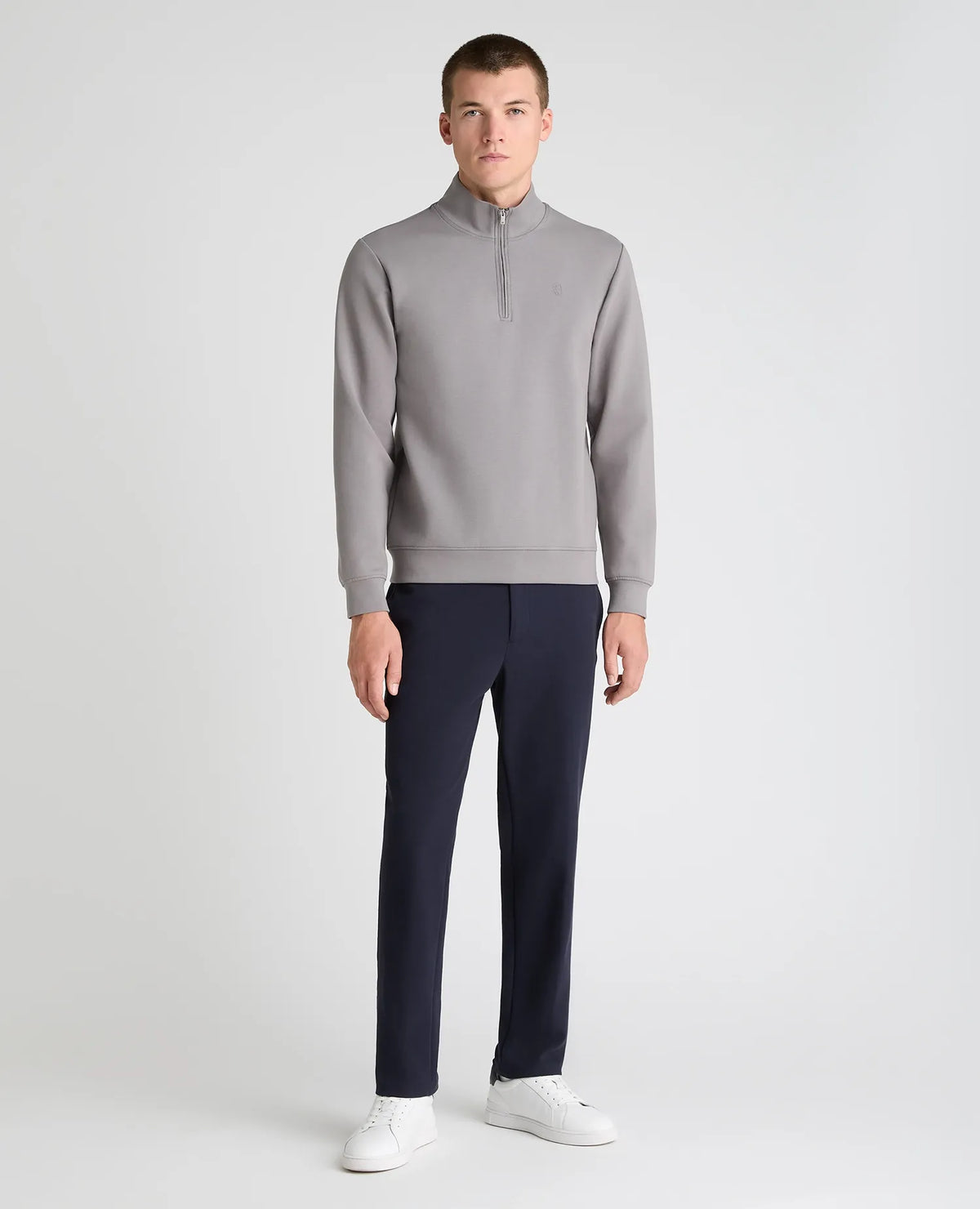 A male model stands against a light gray studio background wearing a light gray quarter-zip pullover, dark navy trousers, and white sneakers.