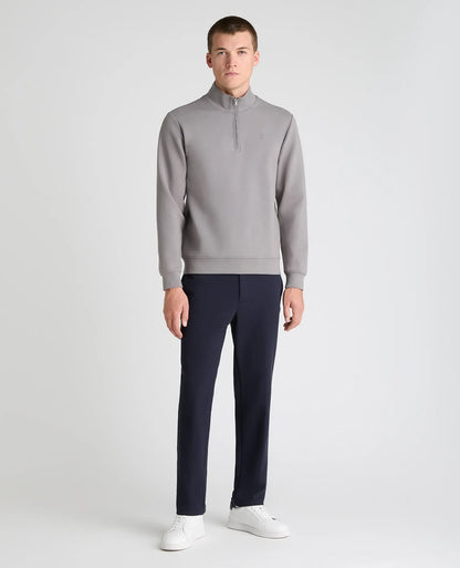 A male model stands against a light gray studio background wearing a light gray quarter-zip pullover, dark navy trousers, and white sneakers.