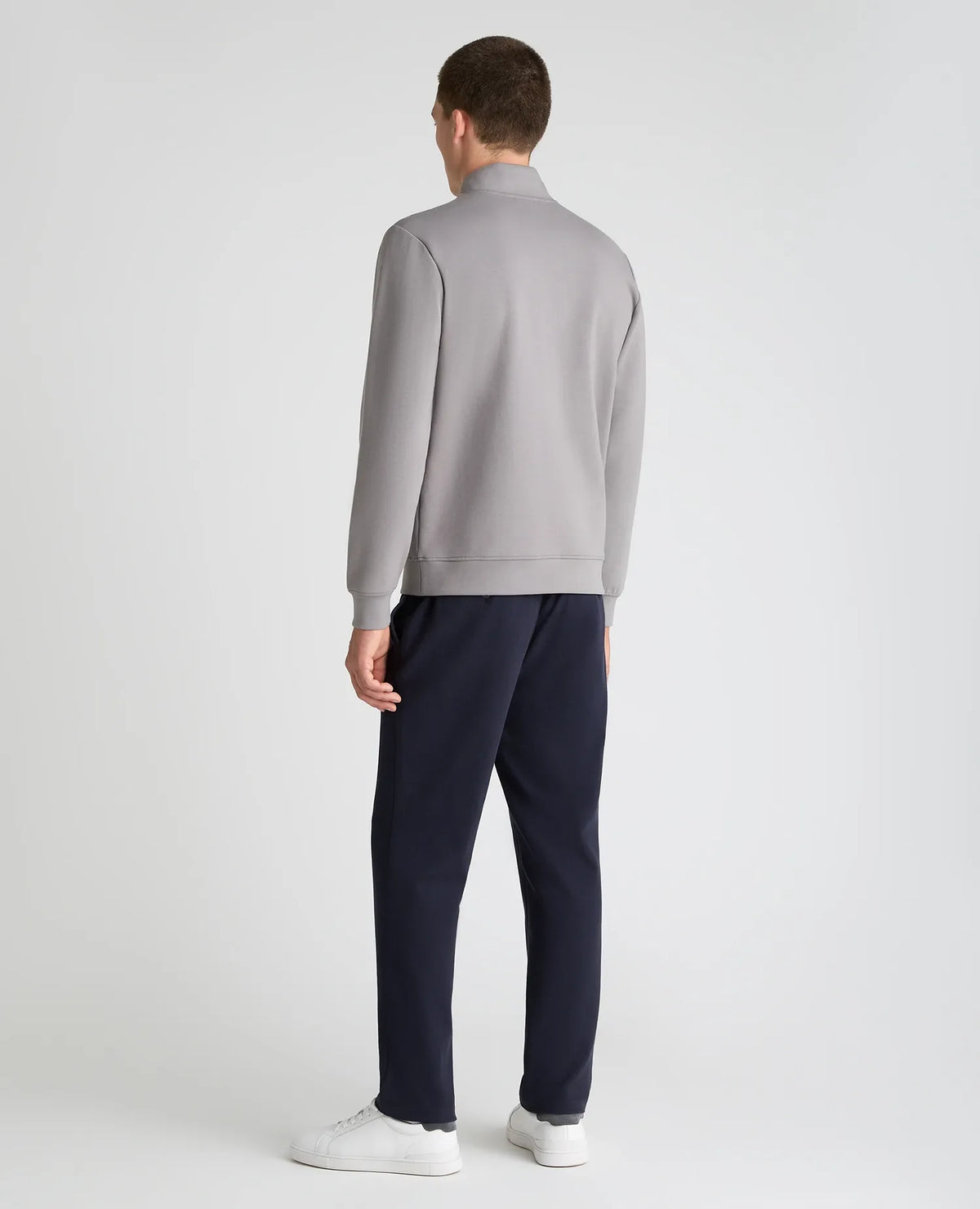 A man viewed from the back wearing a light gray mock-neck sweatshirt, navy blue trousers, and white sneakers, standing against a light gray studio background.