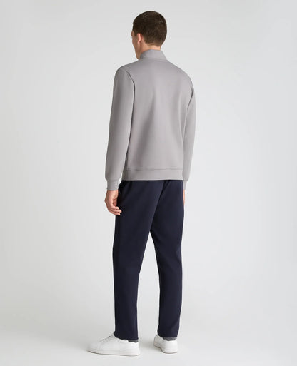 A man viewed from the back wearing a light gray mock-neck sweatshirt, navy blue trousers, and white sneakers, standing against a light gray studio background.