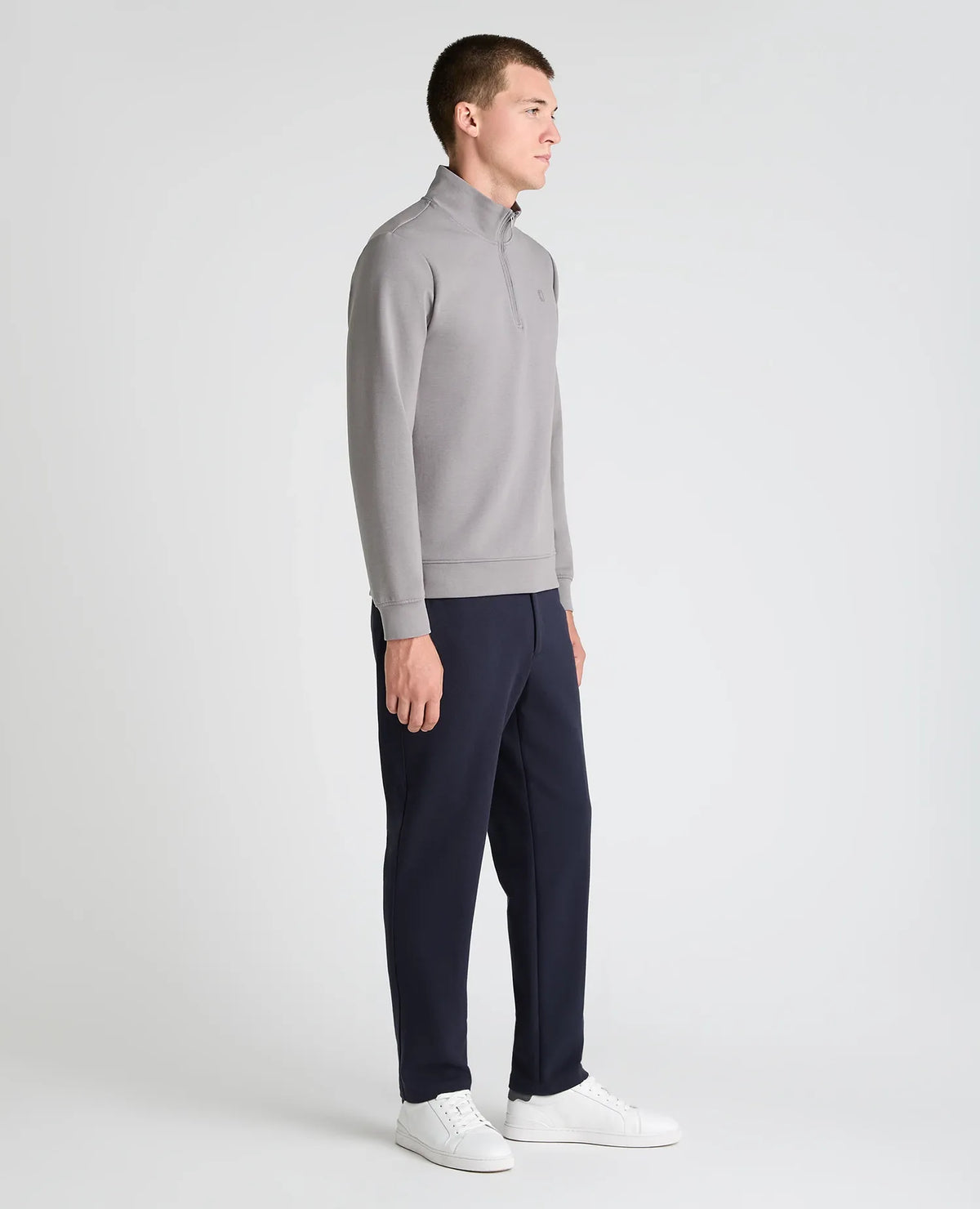 A man in profile wearing a light gray quarter-zip pullover sweater, navy blue pants, and white sneakers, standing against a light gray studio background.