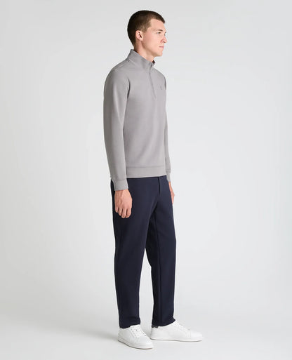 A man in profile wearing a light gray quarter-zip pullover sweater, navy blue pants, and white sneakers, standing against a light gray studio background.