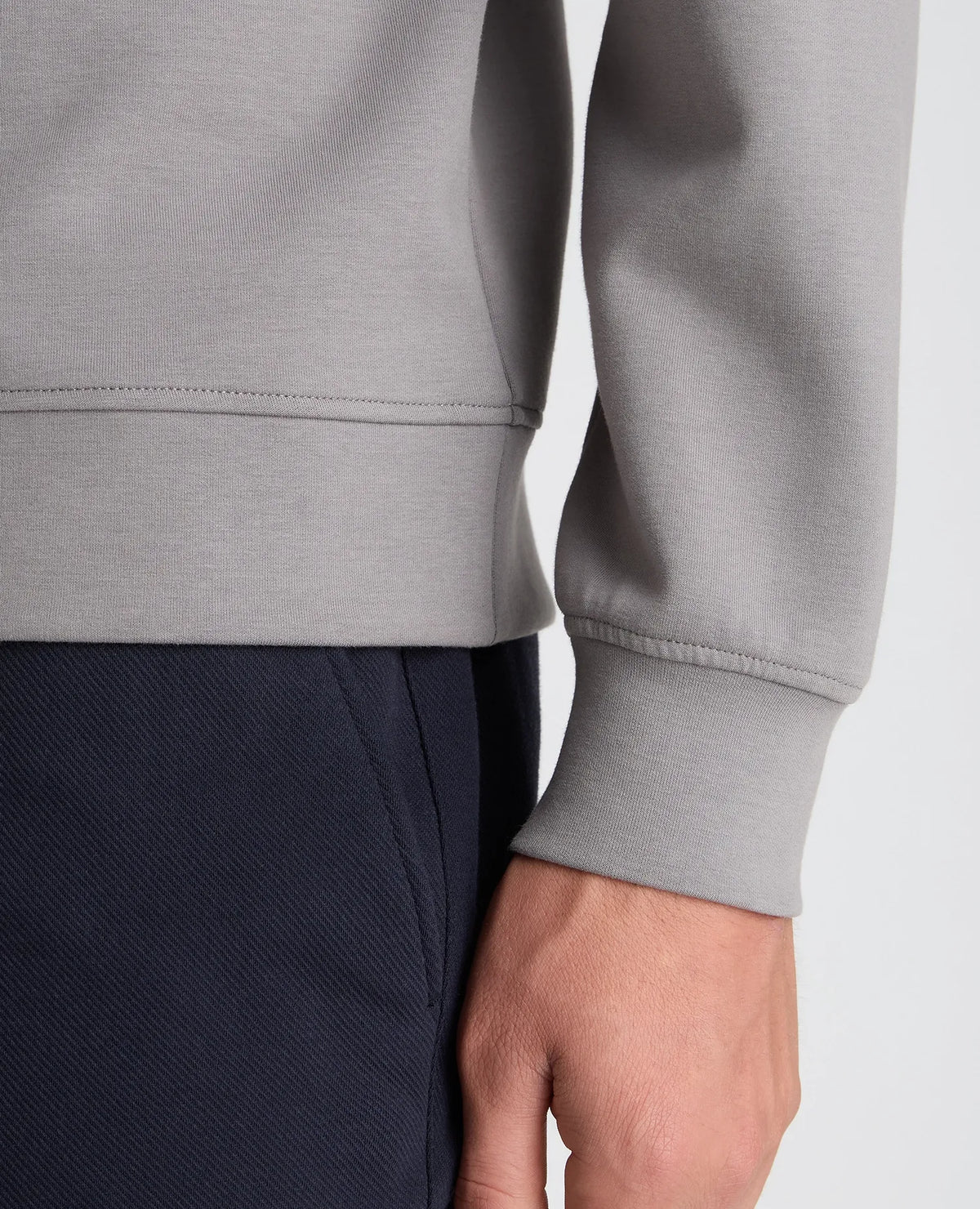Close-up of a person wearing a light gray sweatshirt and dark navy pants, showing the ribbed cuffs and hem of the sweatshirt along with the hand.