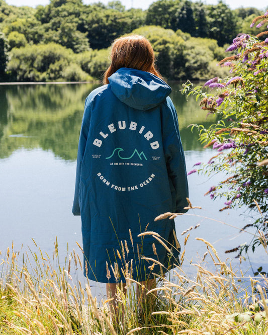 Person wearing a blue 'BLEUBIRD' jacket by a lake with trees and flowers in the background