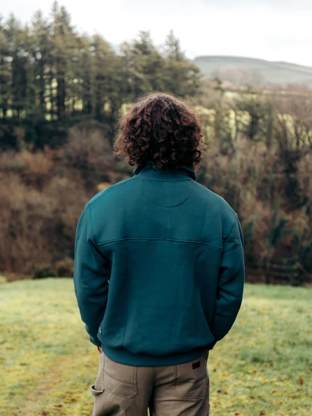Person wearing a teal jacket standing in a field with trees in the background