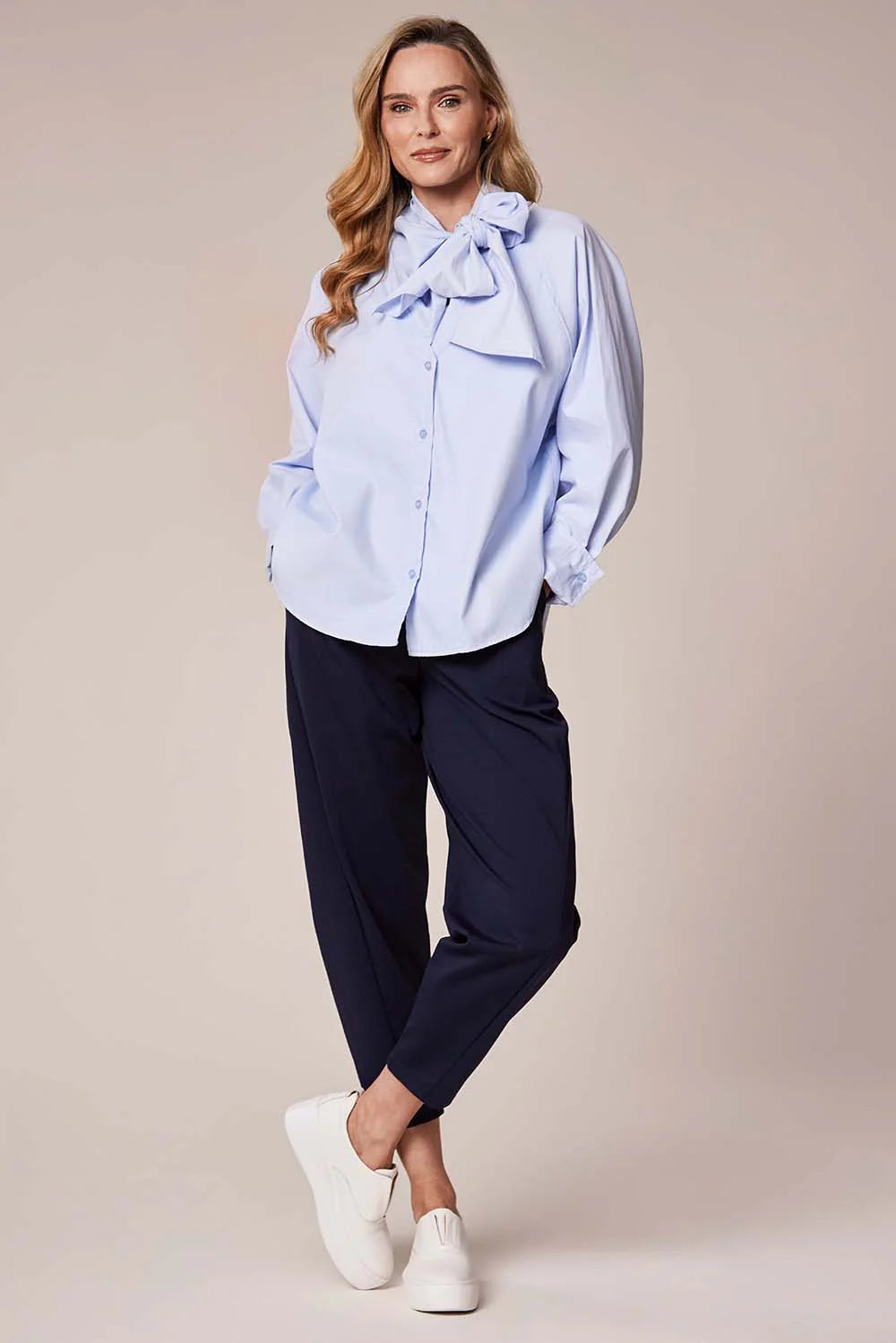 Woman wearing a light blue blouse with a bow detail and dark pants on a beige background