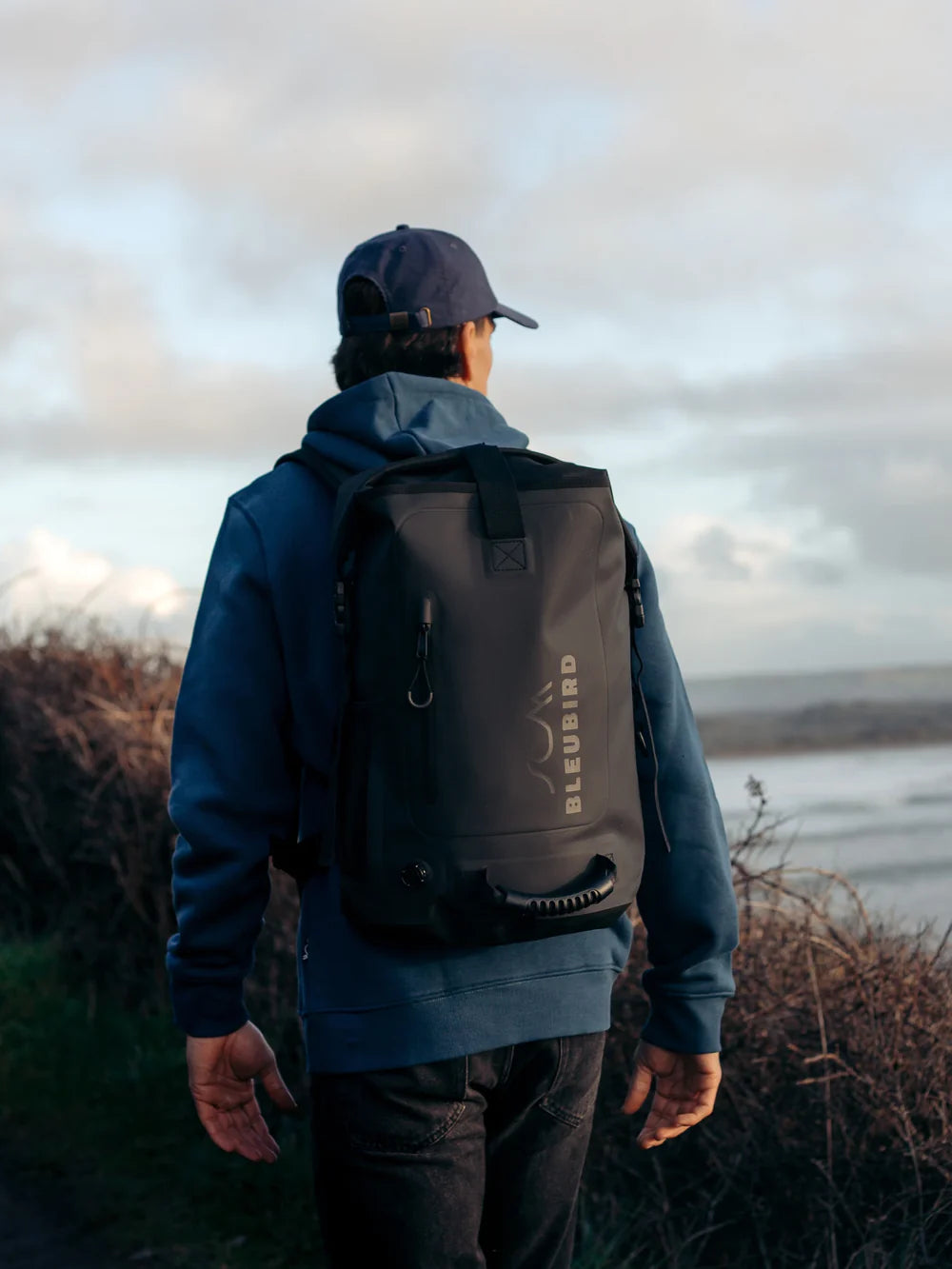 Person wearing a black backpack with 'Bluebird' branding, standing outdoors near water.
