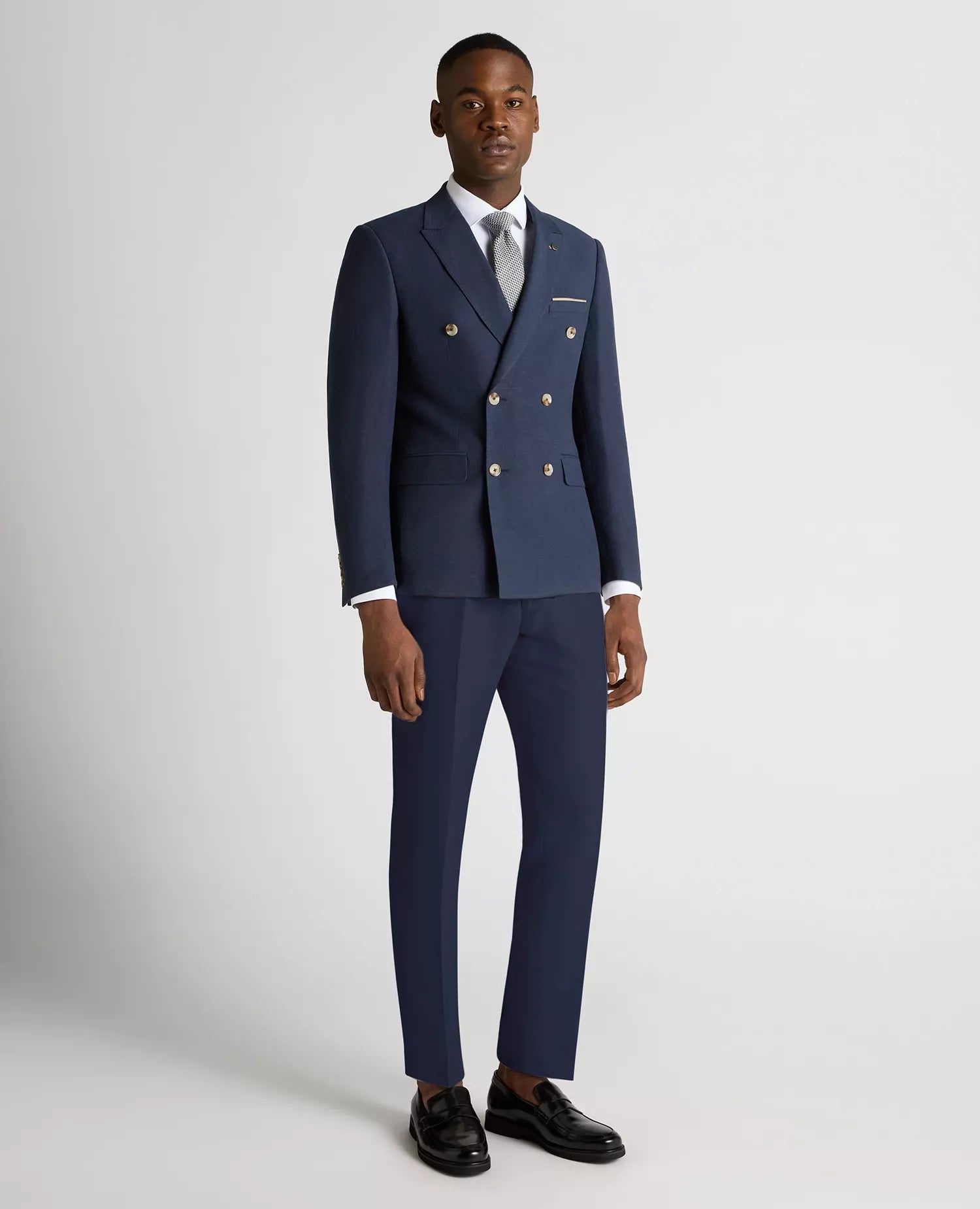 Man wearing a navy blue suit with a white shirt and black shoes on a light gray background