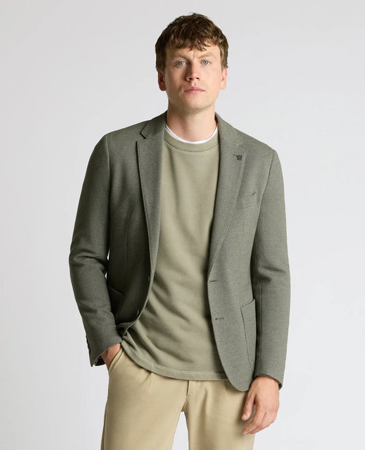 A man wearing a muted sage green blazer over a matching sage green crewneck sweater, paired with beige trousers, photographed against a light gray studio background.