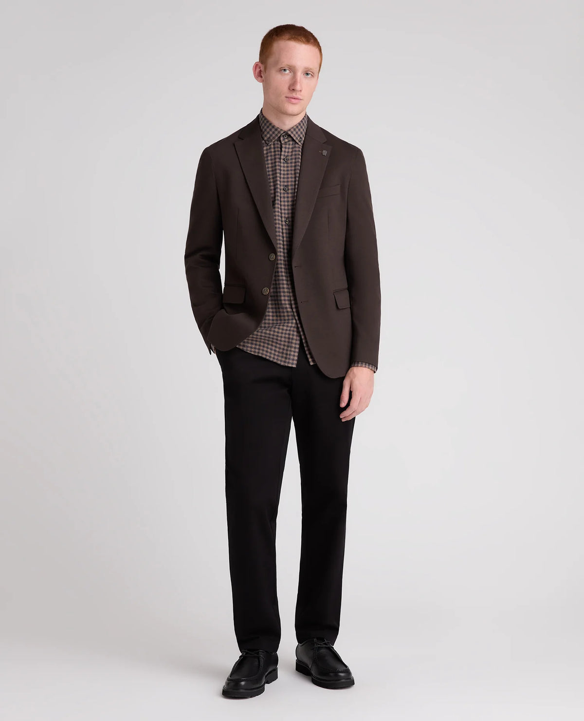 A man modeling a dark brown blazer over a brown and black checkered shirt, paired with black trousers and black shoes, against a light gray studio background.