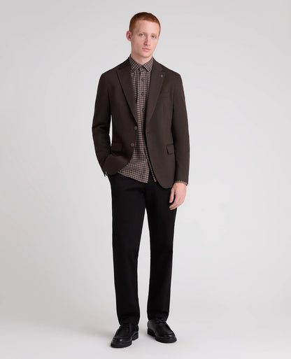 A man modeling a dark brown blazer over a brown and black checkered shirt, paired with black trousers and black shoes, against a light gray studio background.