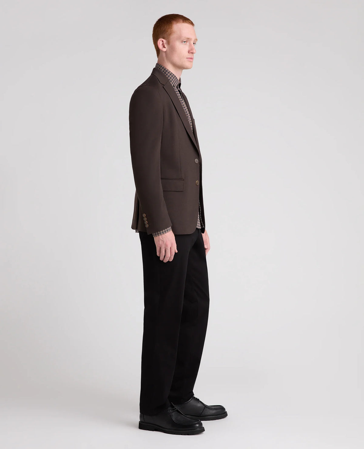 A man in profile wearing a dark brown blazer, black trousers, and black shoes, standing against a light gray studio background.
