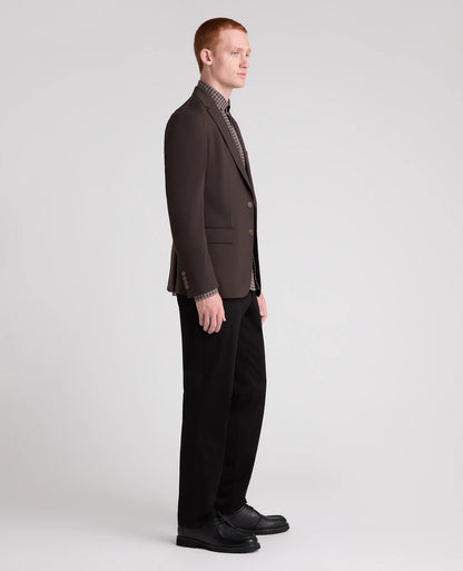 A man in profile wearing a dark brown blazer, black trousers, and black shoes, standing against a light gray studio background.