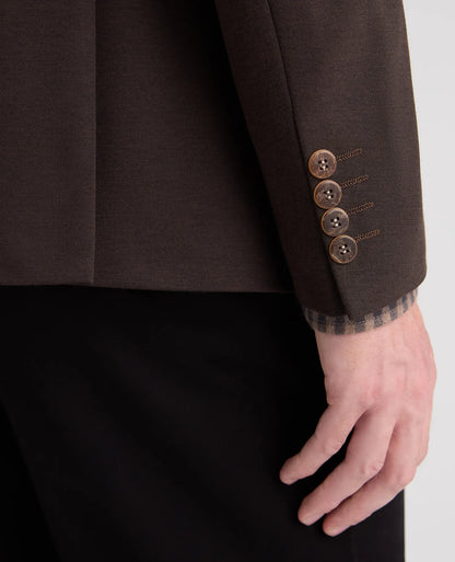 Close-up of a dark brown tailored jacket sleeve with four bronze buttons and a plaid lining at the cuff, worn with black trousers. A hand is visible at the wrist.
