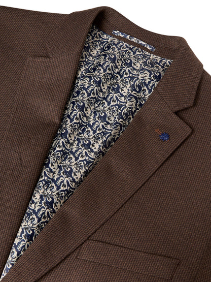 Close-up of a brown textured blazer jacket with a patterned navy and beige lining visible inside the collar, and a small embroidered logo on the lapel.