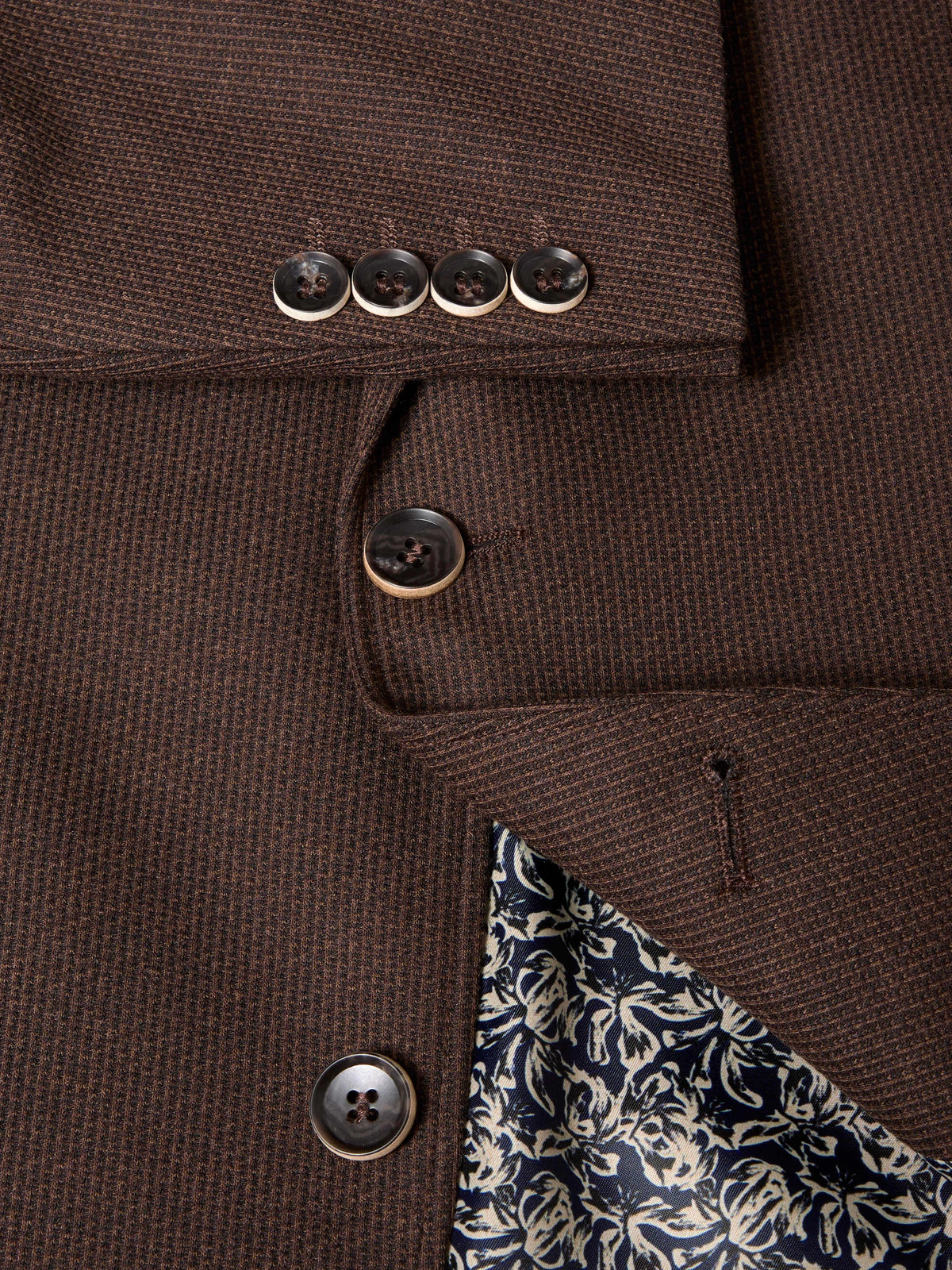 Close-up of a brown textured wool-blend blazer showing the buttoned cuff with four buttons, the front button closure, and a patterned navy and cream lining.
