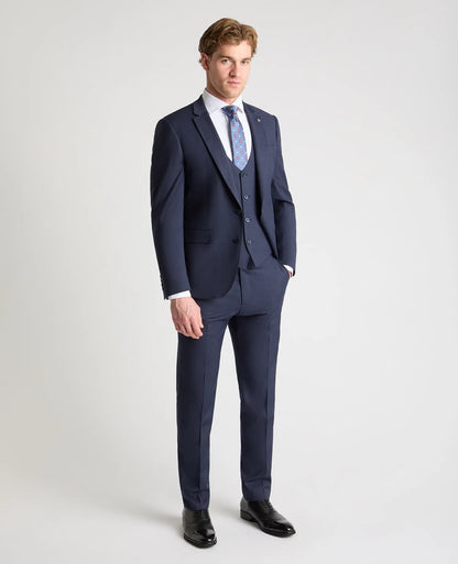 A man modeling a navy blue three-piece suit with a white dress shirt, a light blue patterned tie, and black dress shoes, standing against a light gray studio background.