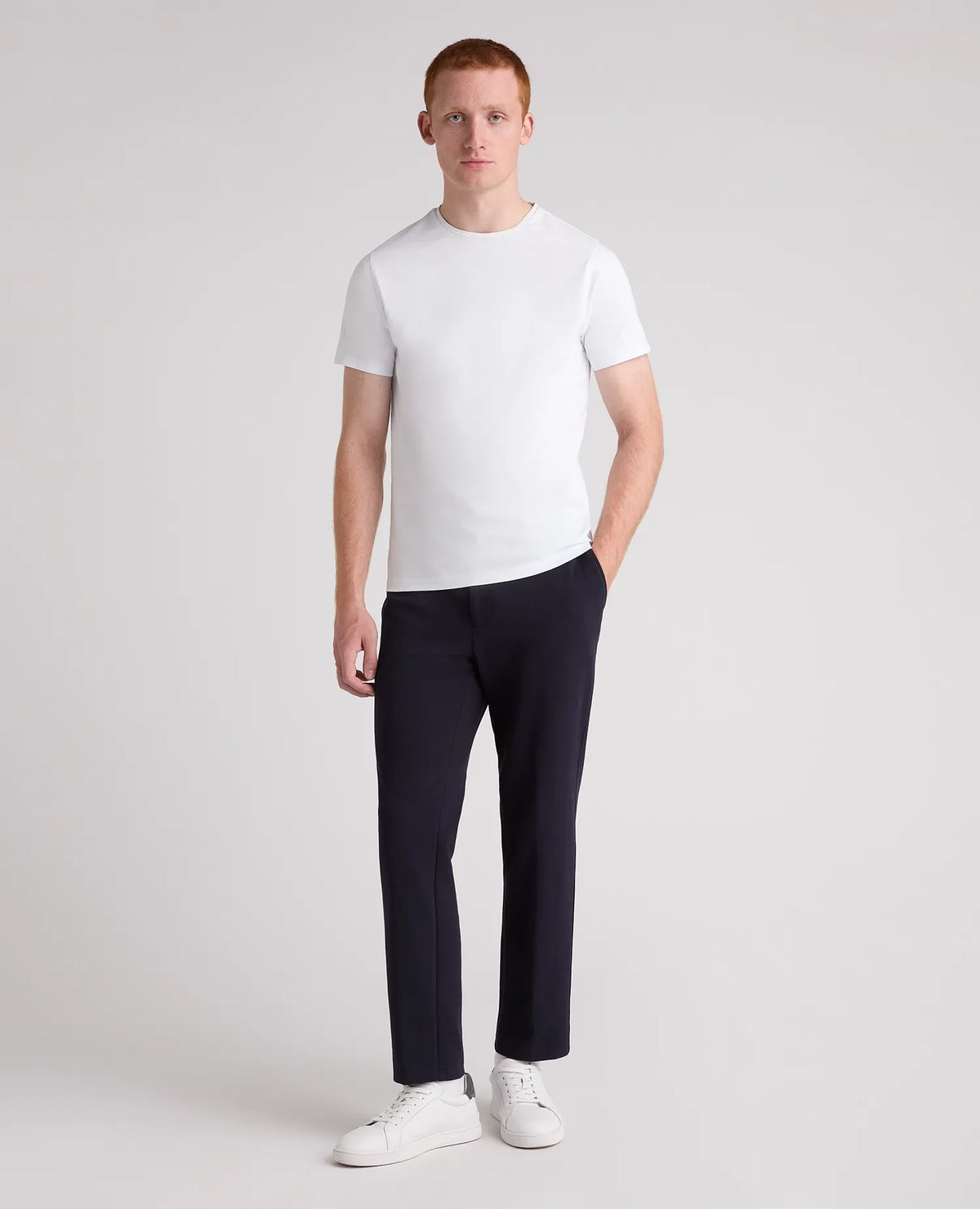 A man modeling a white short-sleeve crewneck t-shirt, dark navy trousers, and white sneakers against a light gray studio background.
