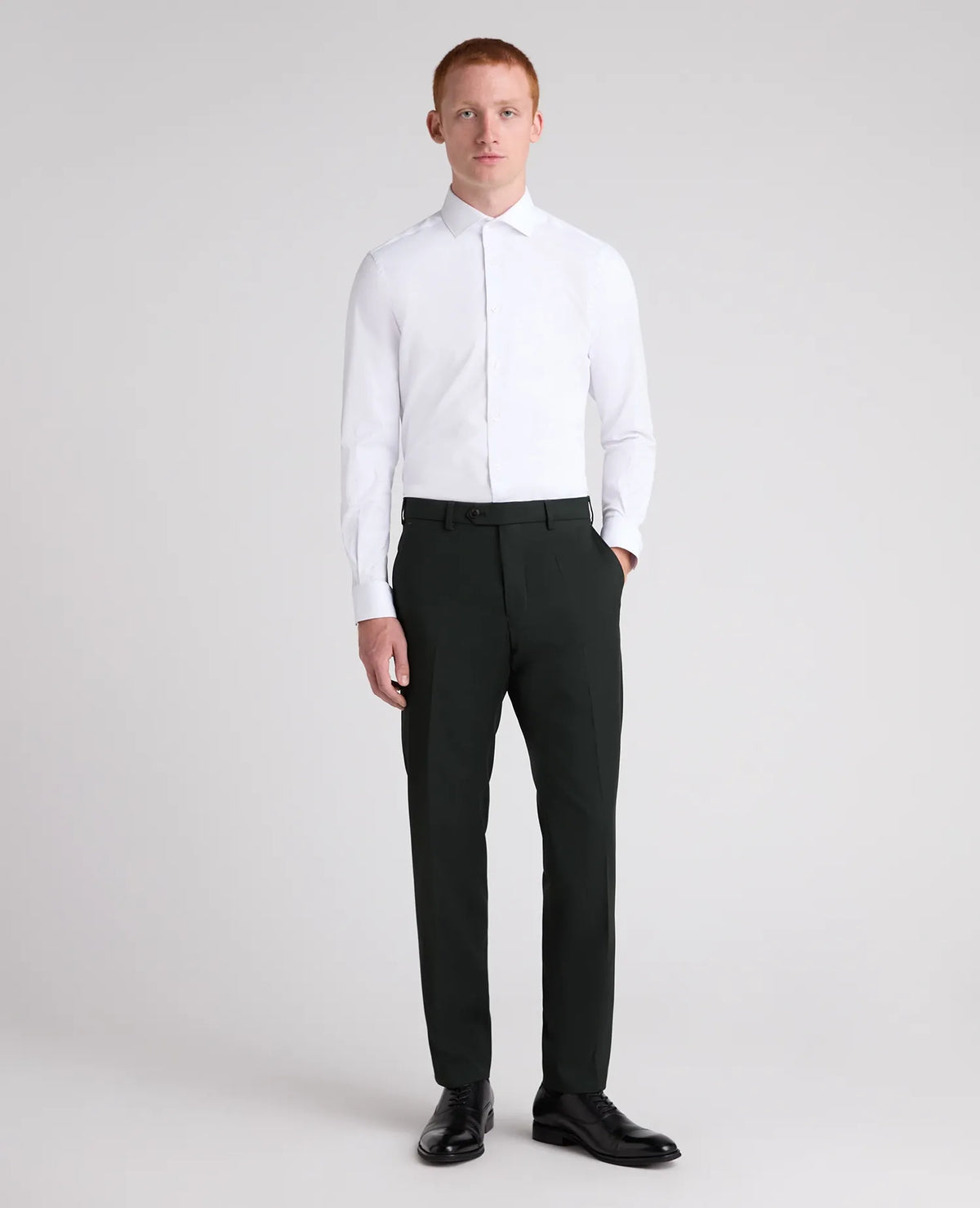 A man modeling a white long-sleeve dress shirt and dark charcoal gray trousers with black dress shoes, standing against a light gray studio background.