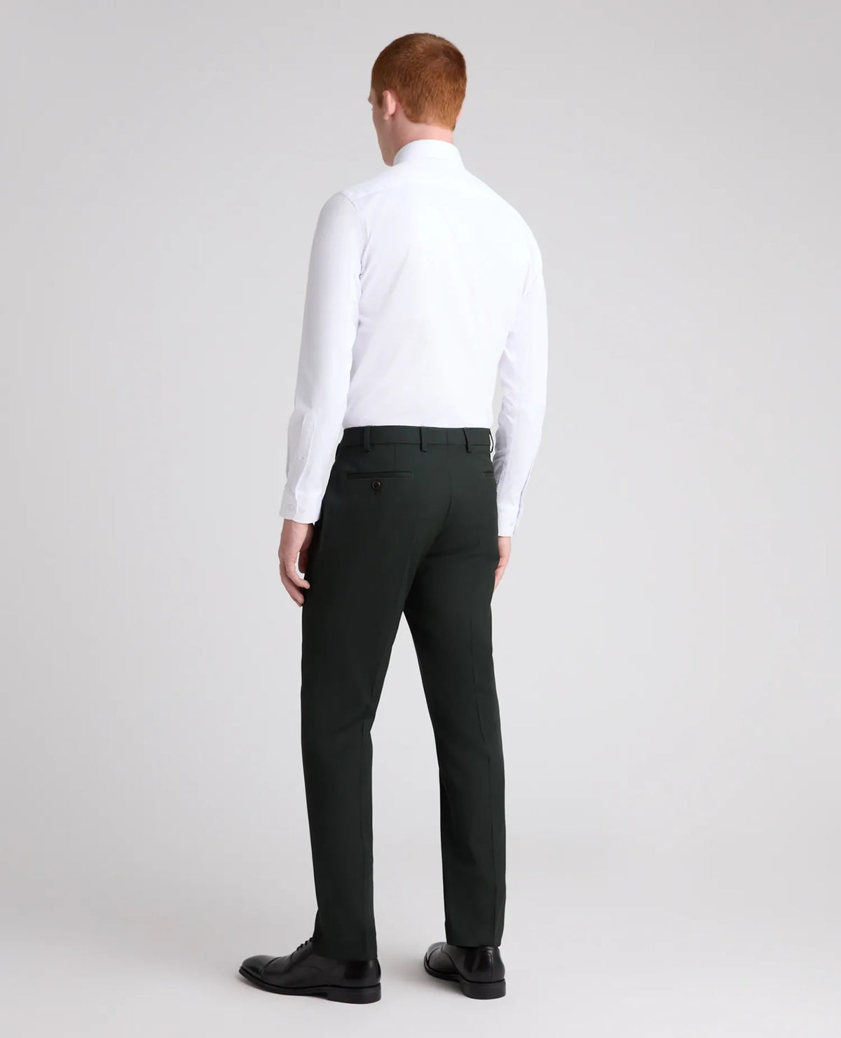 A man viewed from the back wearing a white long-sleeve dress shirt, dark charcoal gray trousers, and black dress shoes, standing against a light gray studio background.