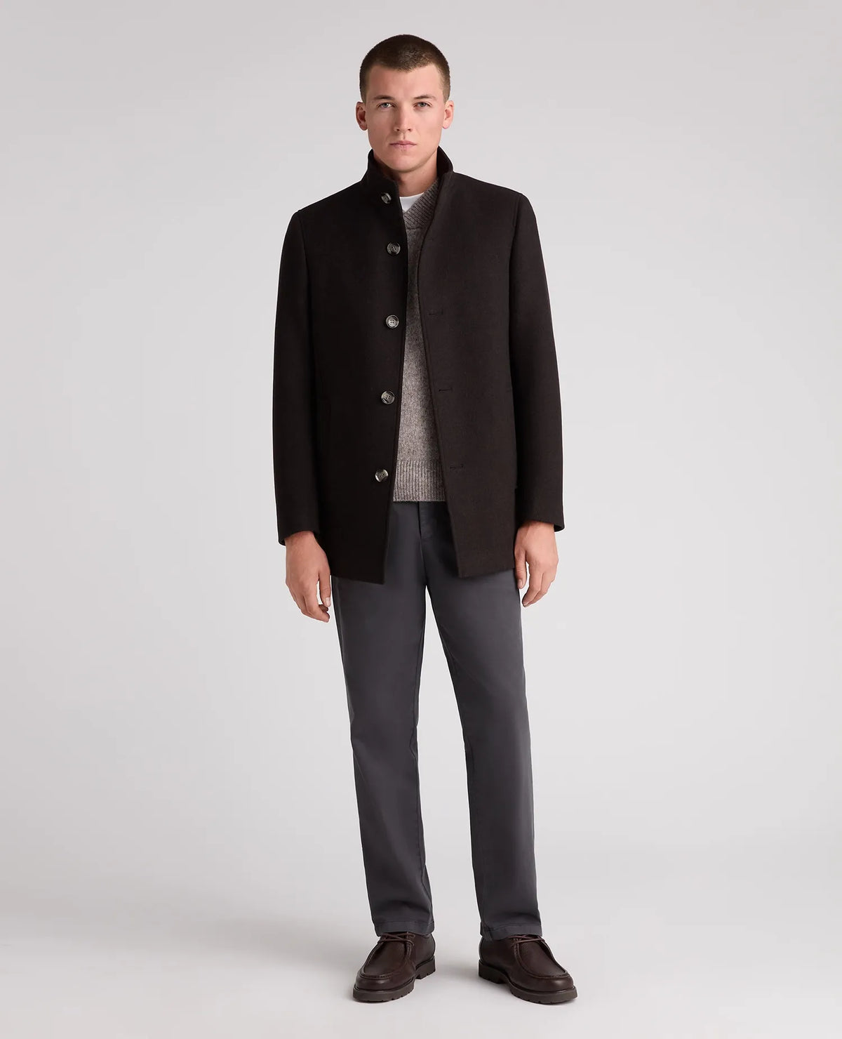 A man modeling a dark brown wool-blend coat with a stand collar, worn over a gray knit sweater, dark gray trousers, and brown leather loafers, shot against a light gray studio background.