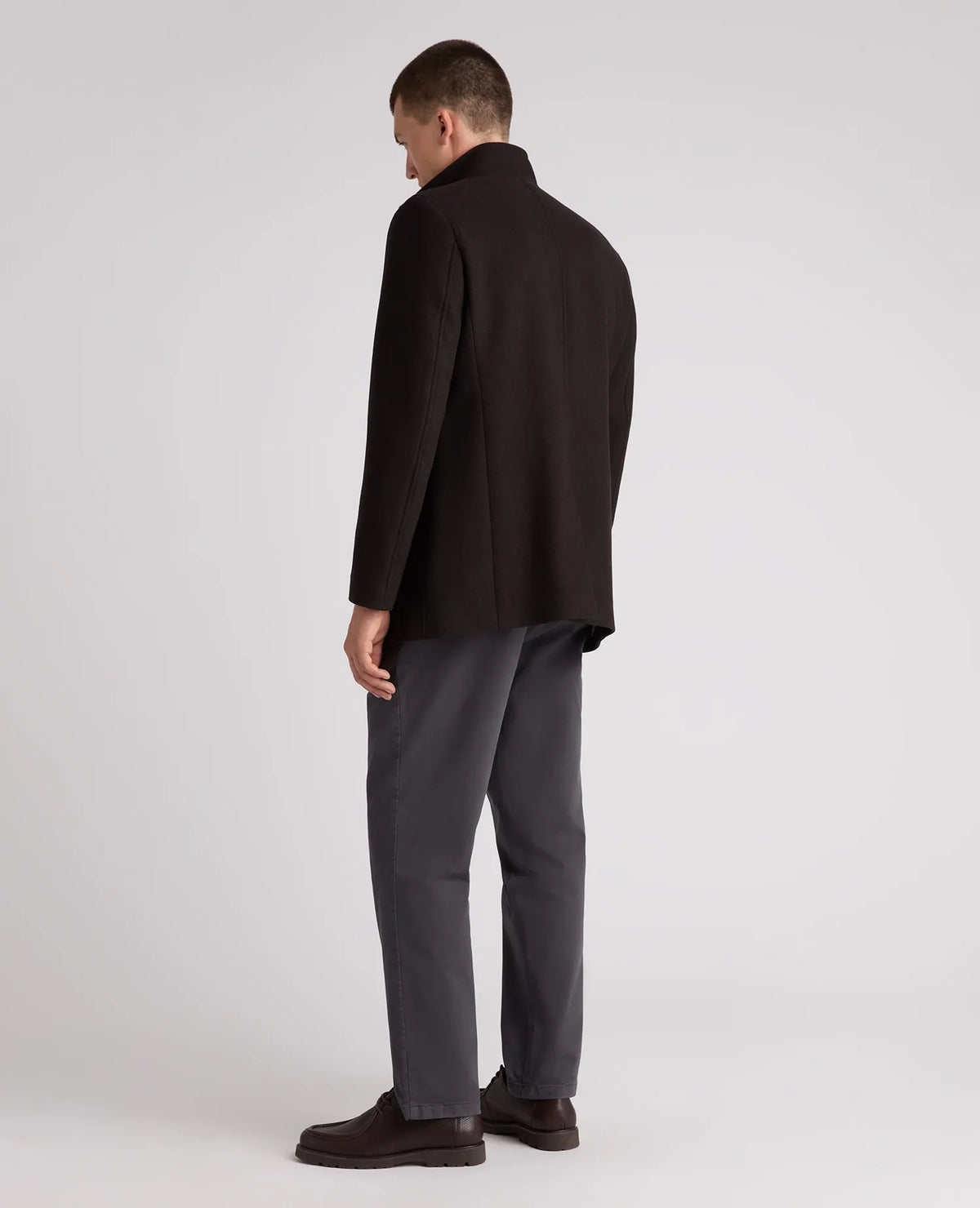 A man shown from the back wearing a dark brown blazer, dark gray trousers, and dark brown leather shoes, standing against a light gray studio background.