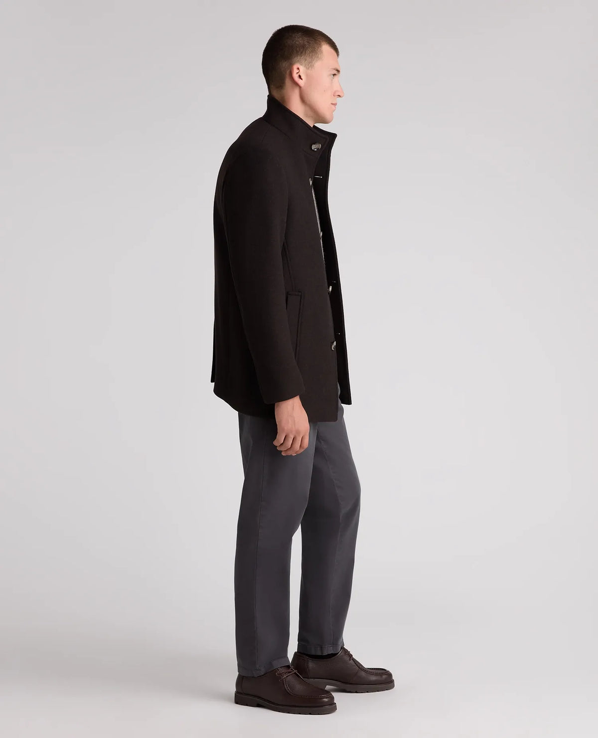 A man in profile wearing a black wool-blend coat, dark gray trousers, and dark brown leather shoes, standing against a light gray studio background.