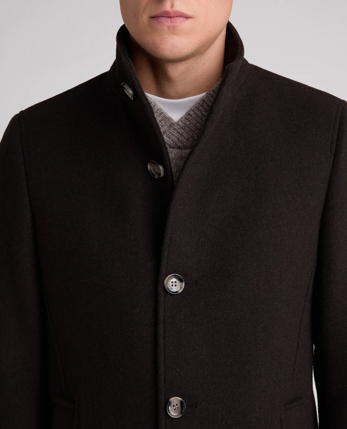 Close-up of a man wearing a dark brown wool-blend coat with a high stand collar, three front buttons, and a gray knit sweater underneath, against a light gray studio background.