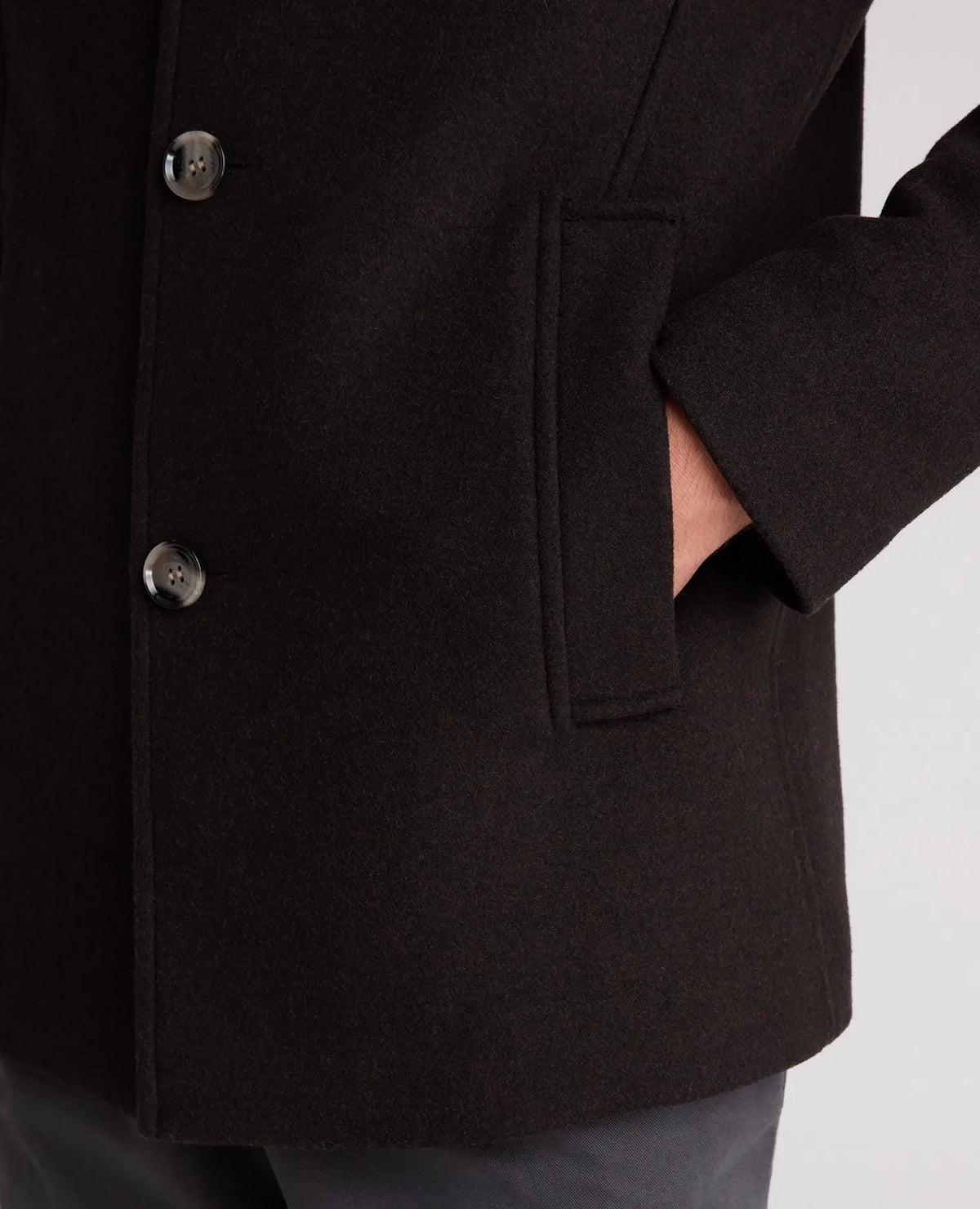 Close-up of a dark brown wool-blend coat with a double-breasted button closure and a side pocket, worn by a model whose hand is in the pocket.
