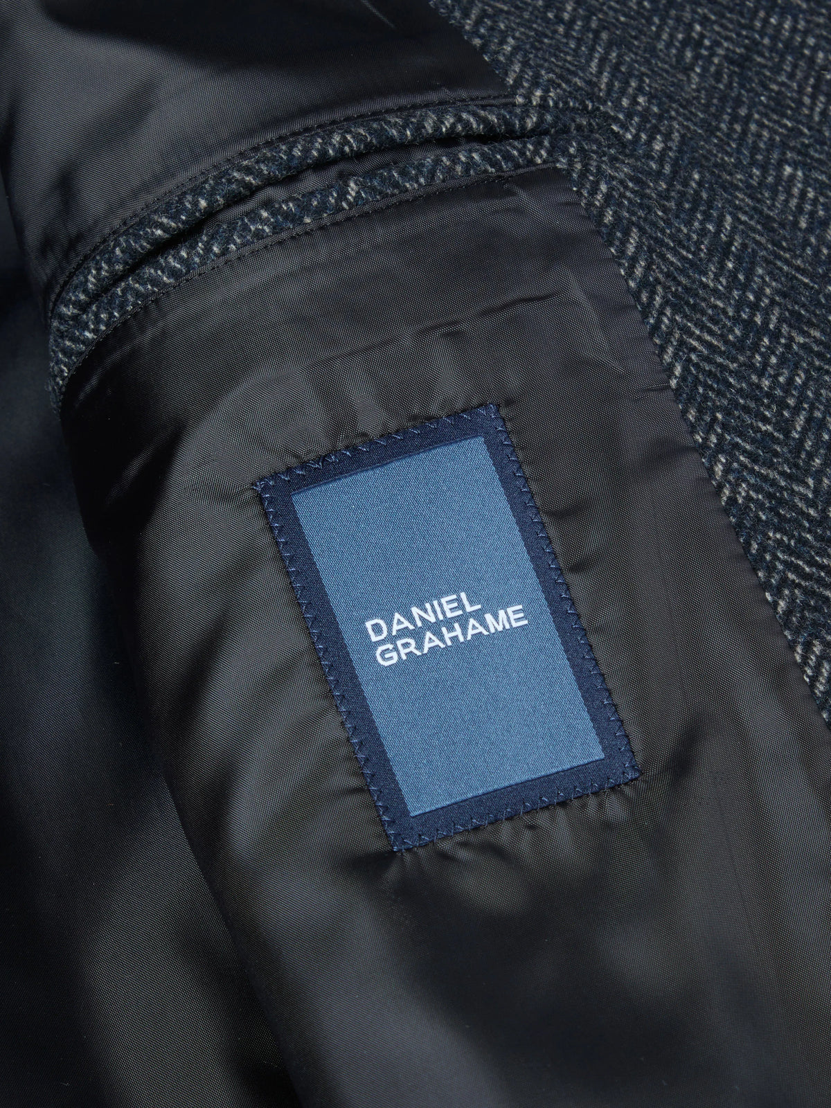Close-up of a Daniel Grahame jacket showing a dark navy herringbone wool fabric and a black nylon lining with a blue brand patch.