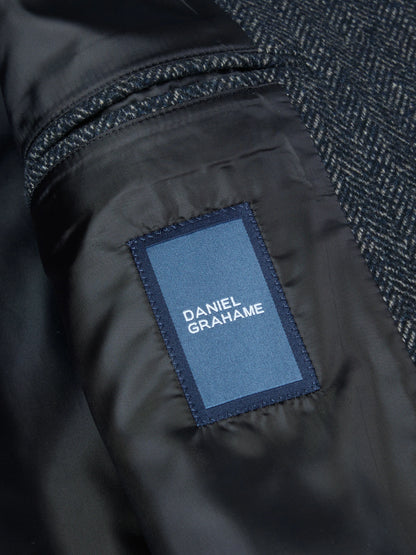 Close-up of a Daniel Grahame jacket showing a dark navy herringbone wool fabric and a black nylon lining with a blue brand patch.