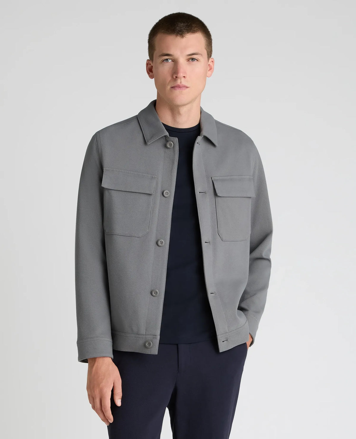 A man wearing a gray utility jacket over a dark navy crewneck shirt and navy trousers, standing against a light gray studio background.