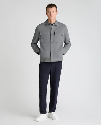 A male model stands against a light gray studio background wearing a gray button-front overshirt with chest pockets, dark navy trousers, and white sneakers.