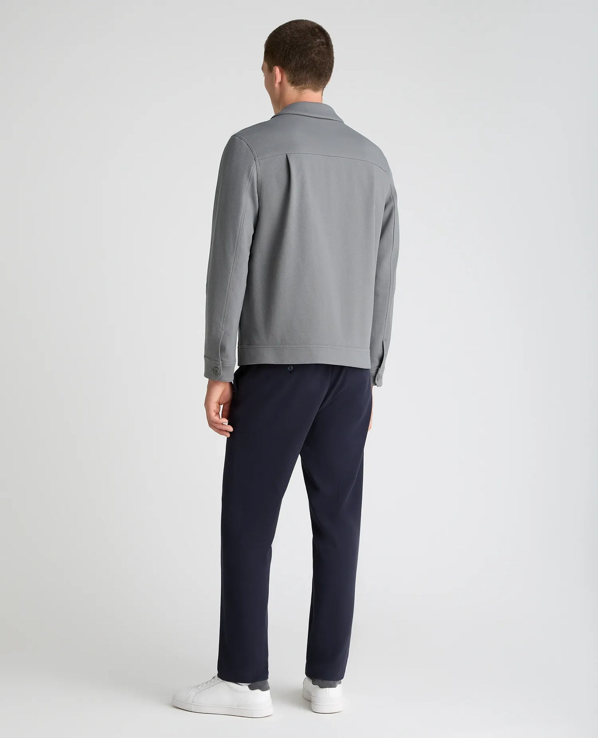 A man viewed from the back wearing a light gray long-sleeve collared shirt, navy blue trousers, and white sneakers, standing against a light gray studio background.