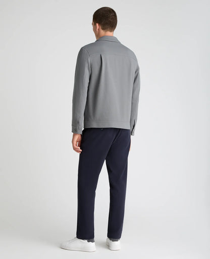 A man viewed from the back wearing a light gray long-sleeve collared shirt, navy blue trousers, and white sneakers, standing against a light gray studio background.