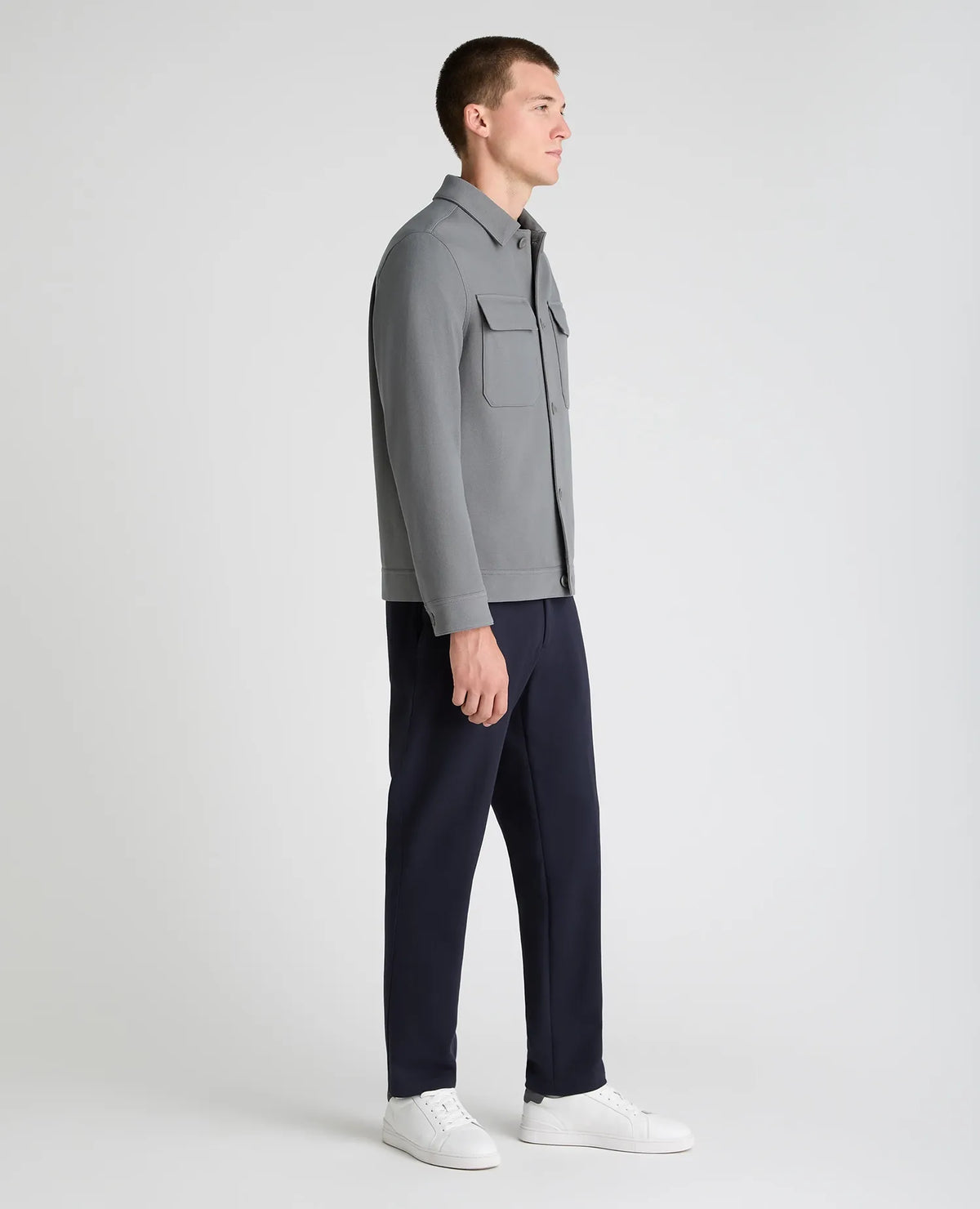 A man in profile wearing a gray long-sleeve button-up shirt, dark navy trousers, and white sneakers, standing against a light gray studio background.