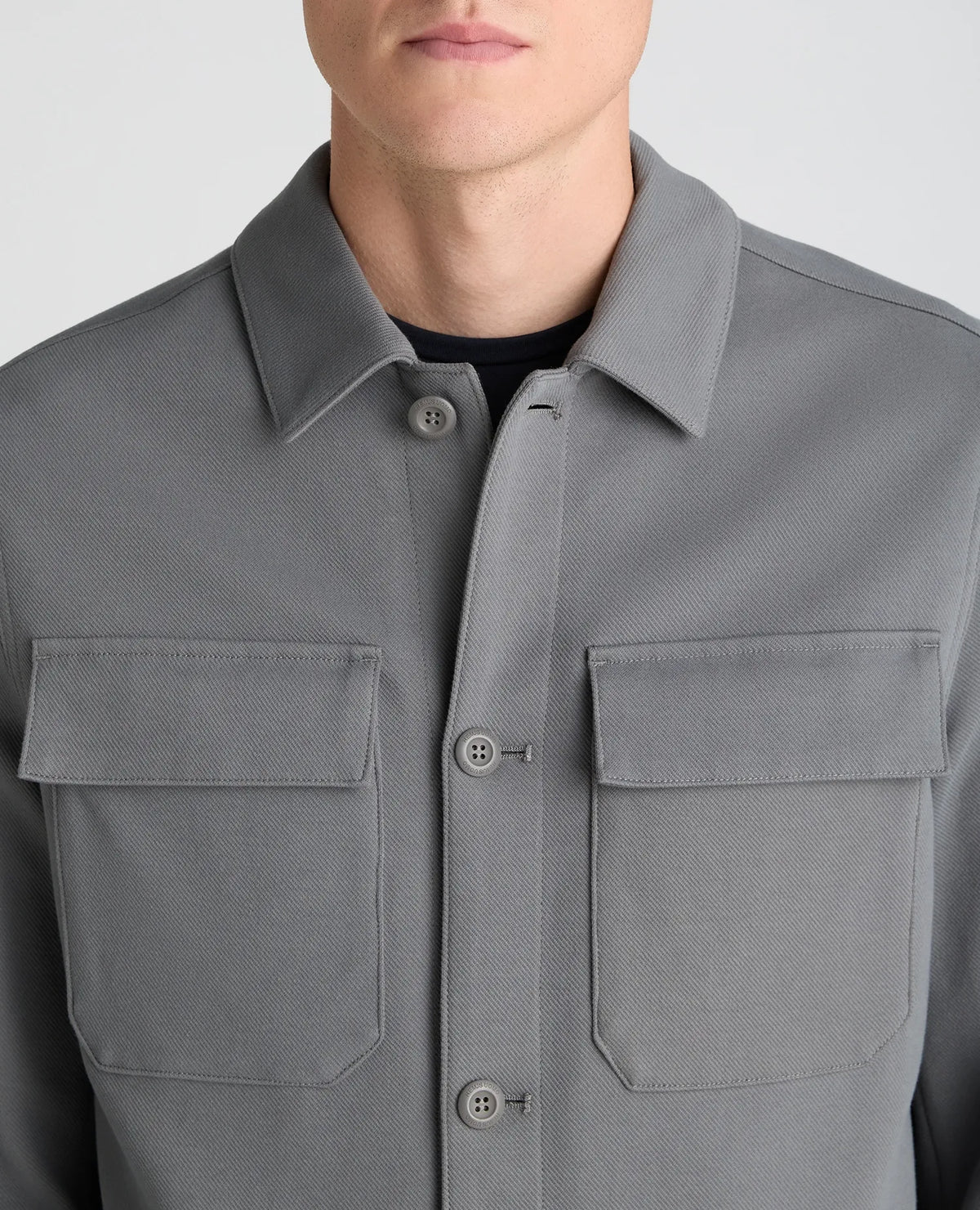 A close-up of a person wearing a gray button-front overshirt or shirt jacket with a pointed collar and two flap chest pockets, photographed against a light gray studio background.