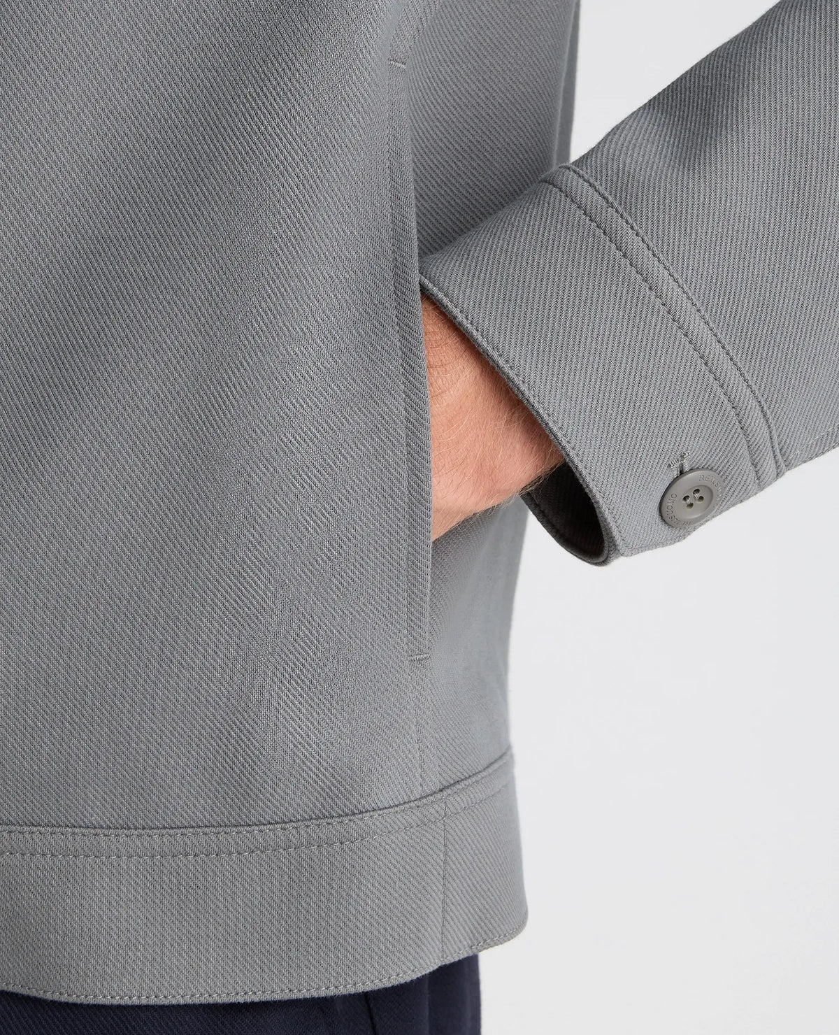 Close-up of a person wearing a gray textured jacket, showing the sleeve with a button cuff and a hand tucked into a pocket, against a light gray background.