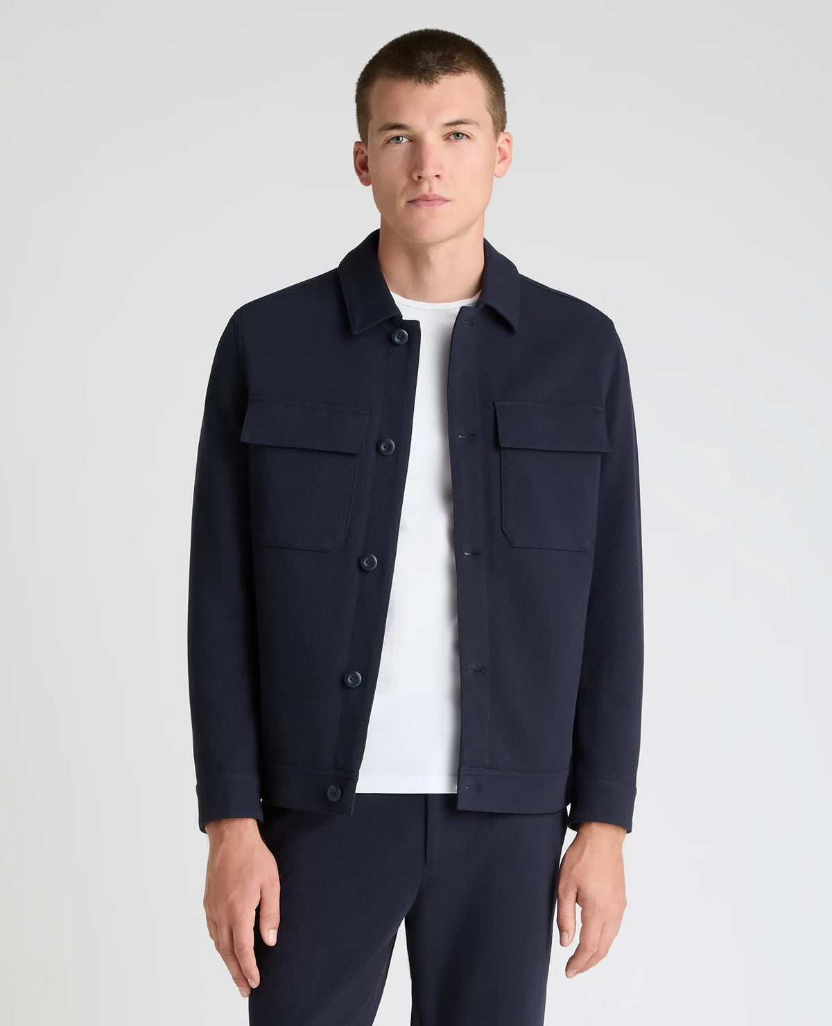 A man wearing a dark navy blue utility jacket over a white t-shirt and matching navy trousers, standing against a light gray studio background.