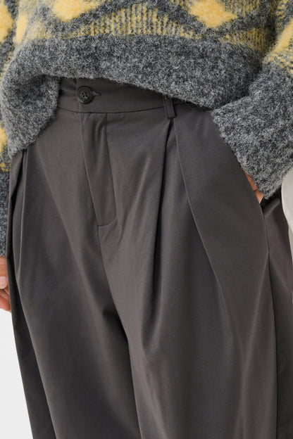 Close-up of a person wearing dark charcoal gray pleated trousers and a fuzzy gray sweater with yellow patterned knit details.