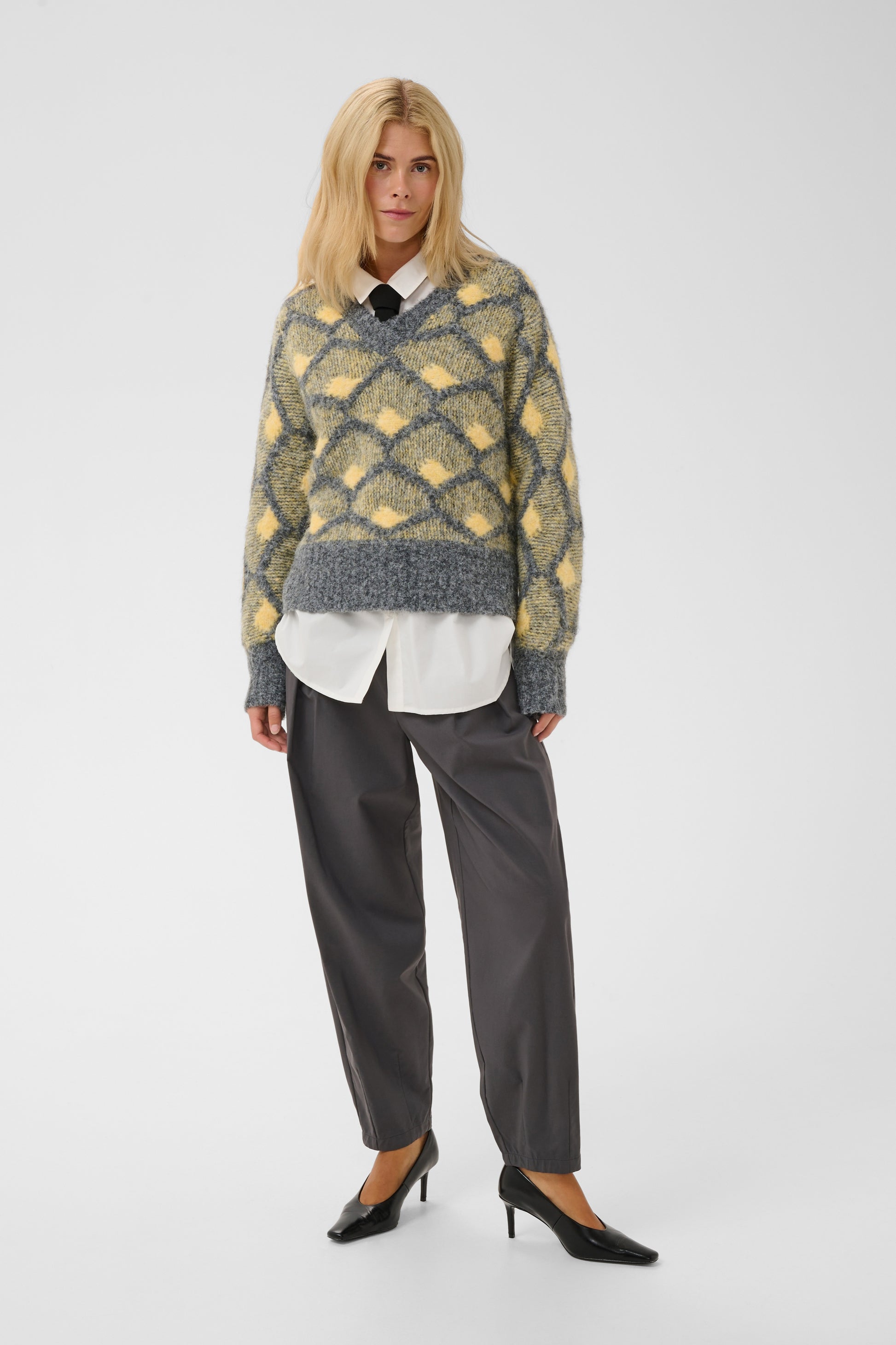 A model stands against a light gray studio background wearing a fuzzy knit V-neck sweater with a yellow and gray diamond pattern, layered over a white collared shirt, dark gray trousers, and black pointed-toe heels.