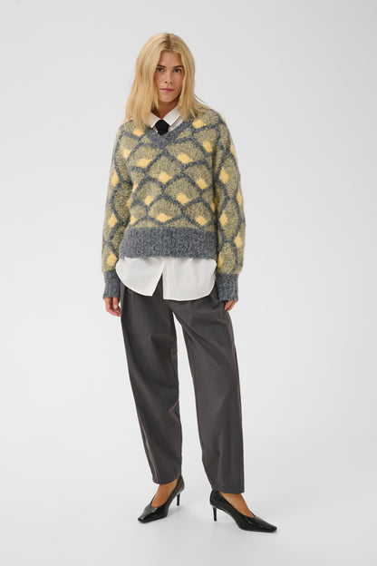 A model stands against a light gray studio background wearing a fuzzy knit V-neck sweater with a yellow and gray diamond pattern, layered over a white collared shirt, dark gray trousers, and black pointed-toe heels.