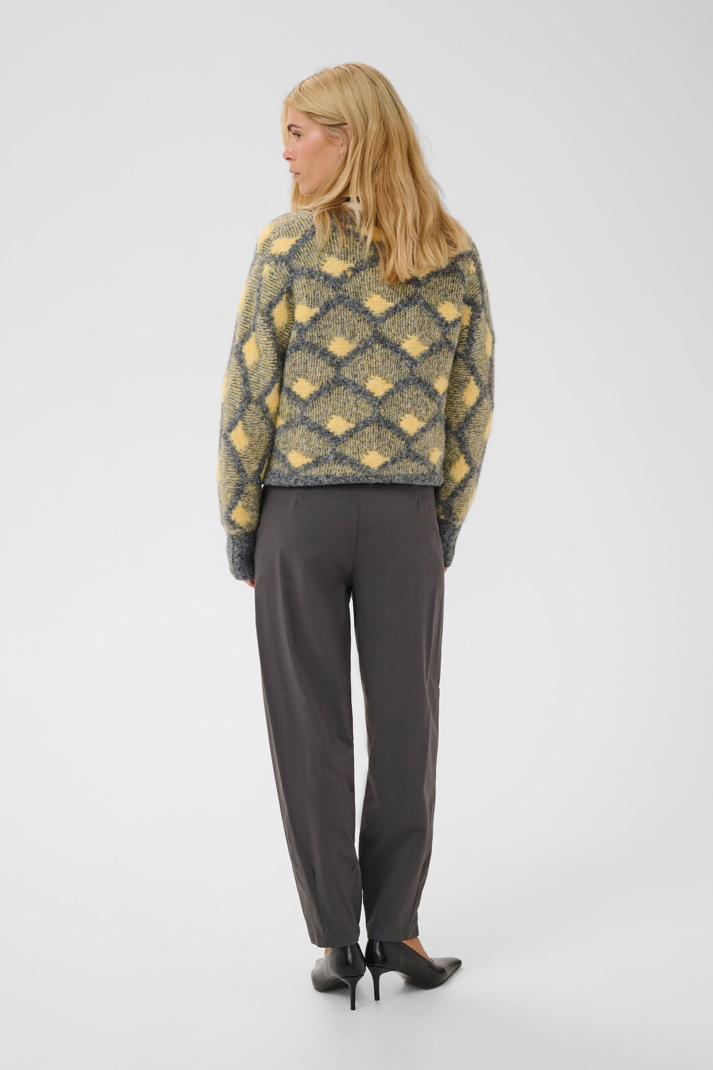 A woman is shown from the back wearing a yellow and gray patterned knit sweater, dark gray trousers, and black heels against a plain light gray studio background.