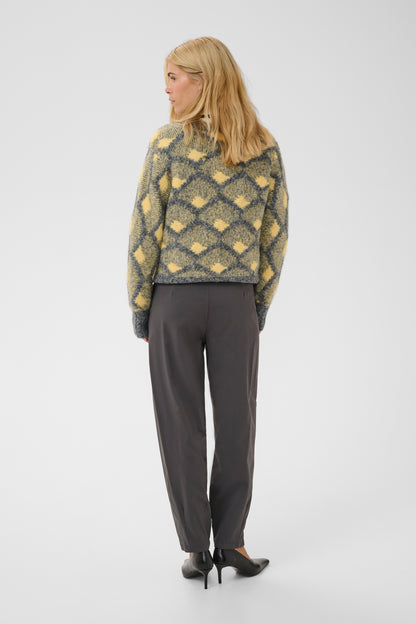 A woman is shown from the back wearing a yellow and gray patterned knit sweater, dark gray trousers, and black heels against a plain light gray studio background.