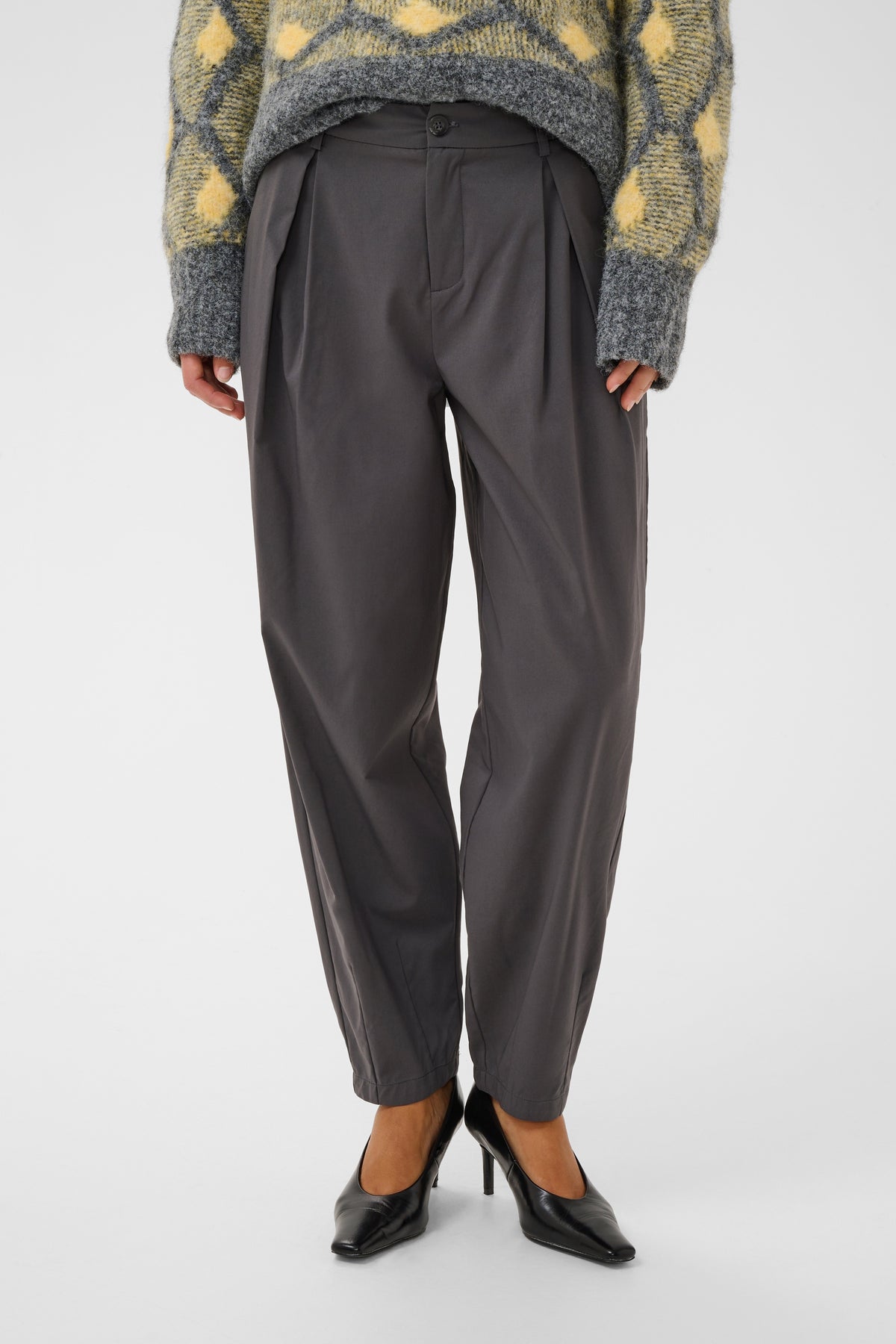 A person wearing dark charcoal gray pleated trousers, a yellow and gray patterned knit sweater, and black pointed-toe heels, shown from the chest down against a light gray studio background.