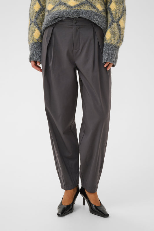 A person wearing dark charcoal gray pleated trousers, a yellow and gray patterned knit sweater, and black pointed-toe heels, shown from the chest down against a light gray studio background.