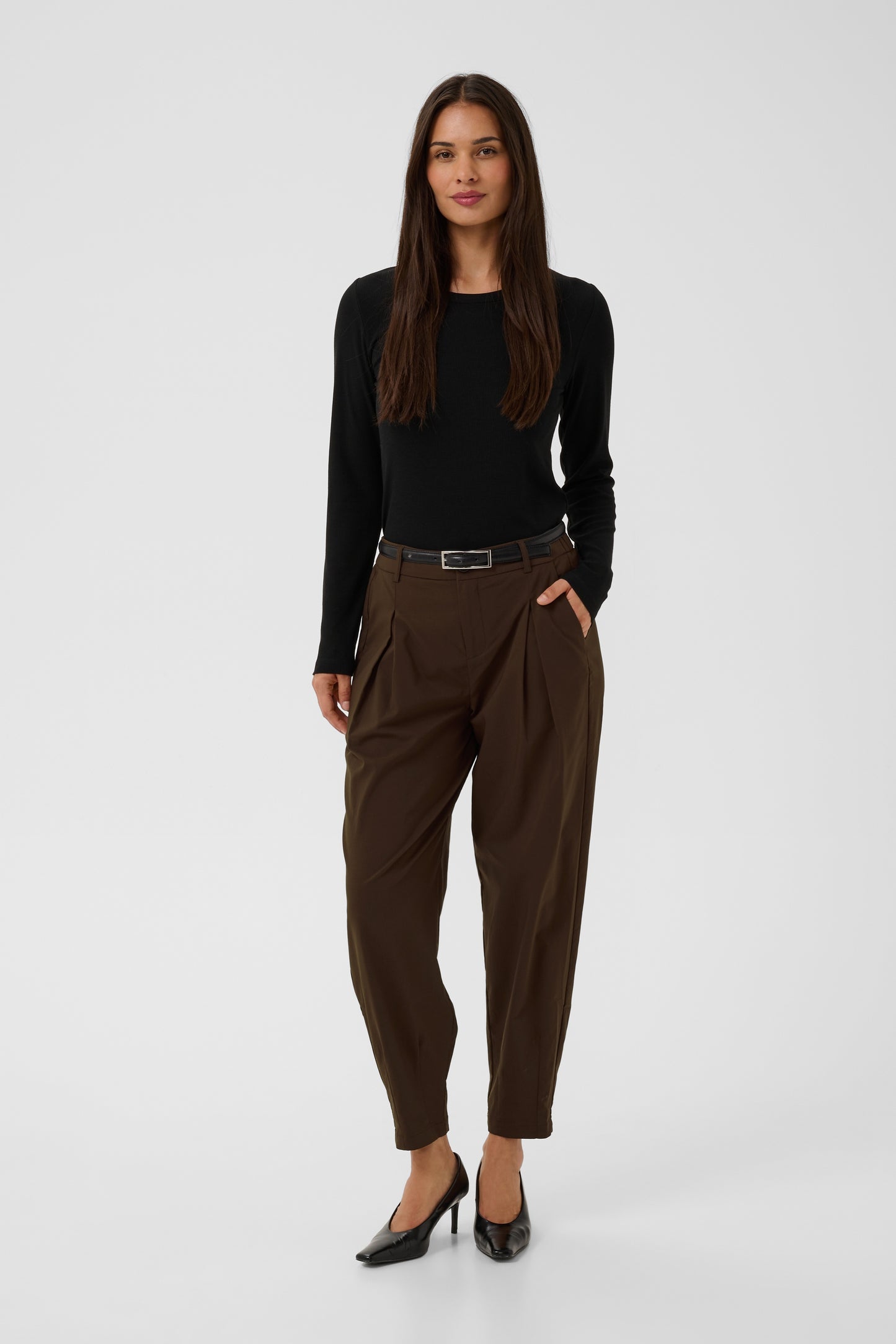A woman modeling a black long-sleeve top and dark brown pleated trousers with a black belt, paired with black heels, against a light gray studio background.