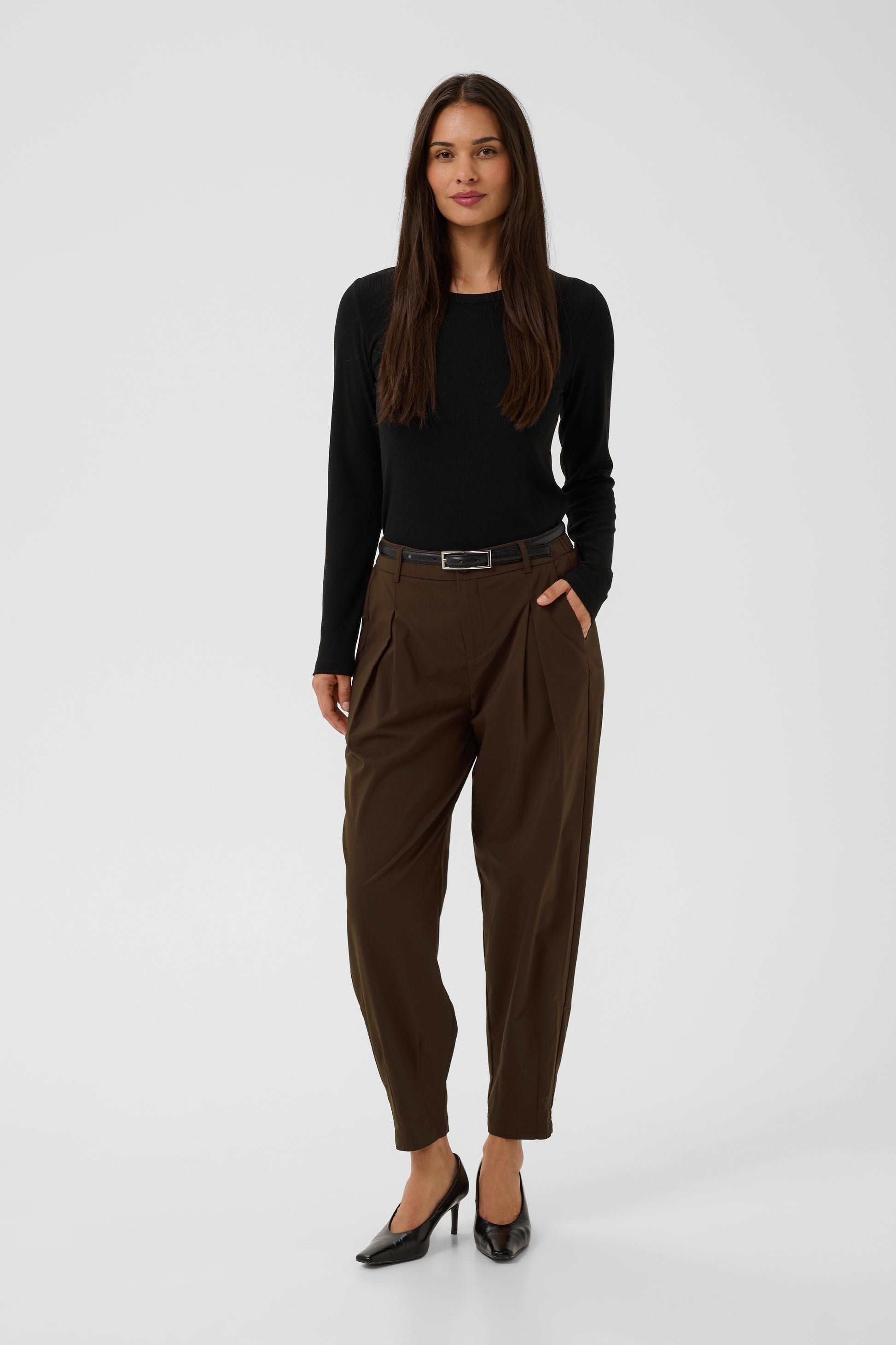A woman modeling a black long-sleeve top and dark brown pleated trousers with a black belt, paired with black heels, against a light gray studio background.